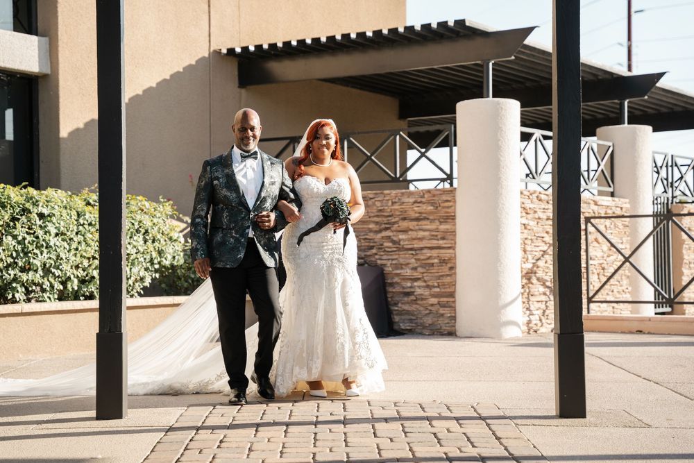 A bride and groom walk together outside a modern venue, she in a white gown, he in a floral tuxedo jacket.