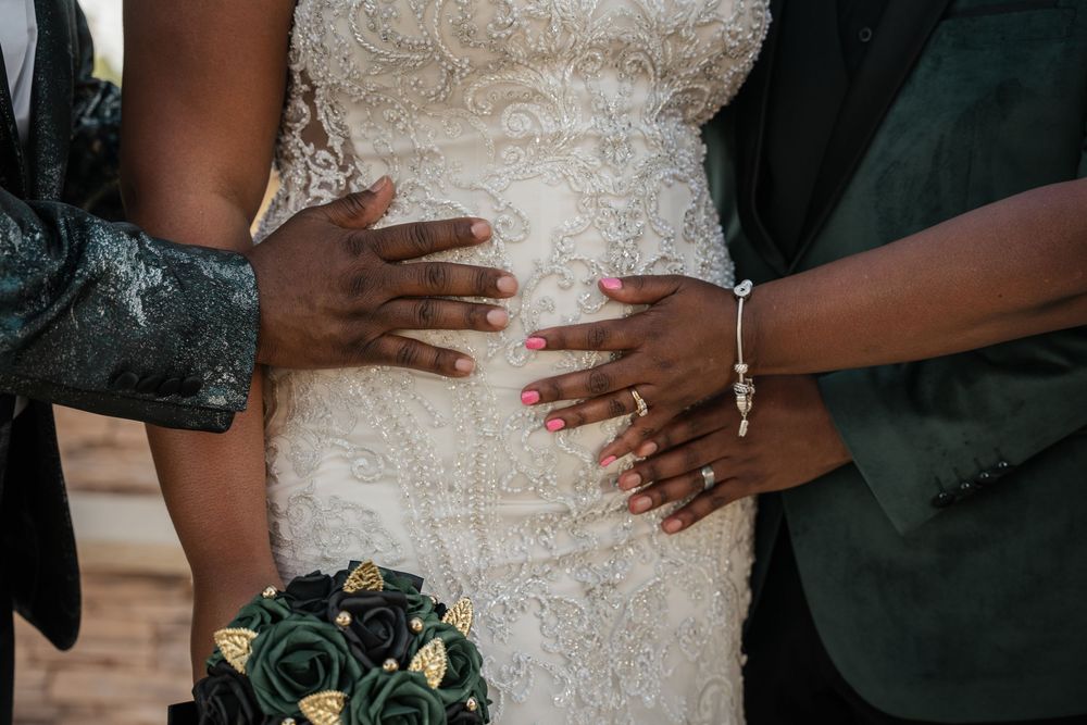 Close-up of hands with pink nails and bracelet on bride in white lace wedding dress holding dark floral bouquet.