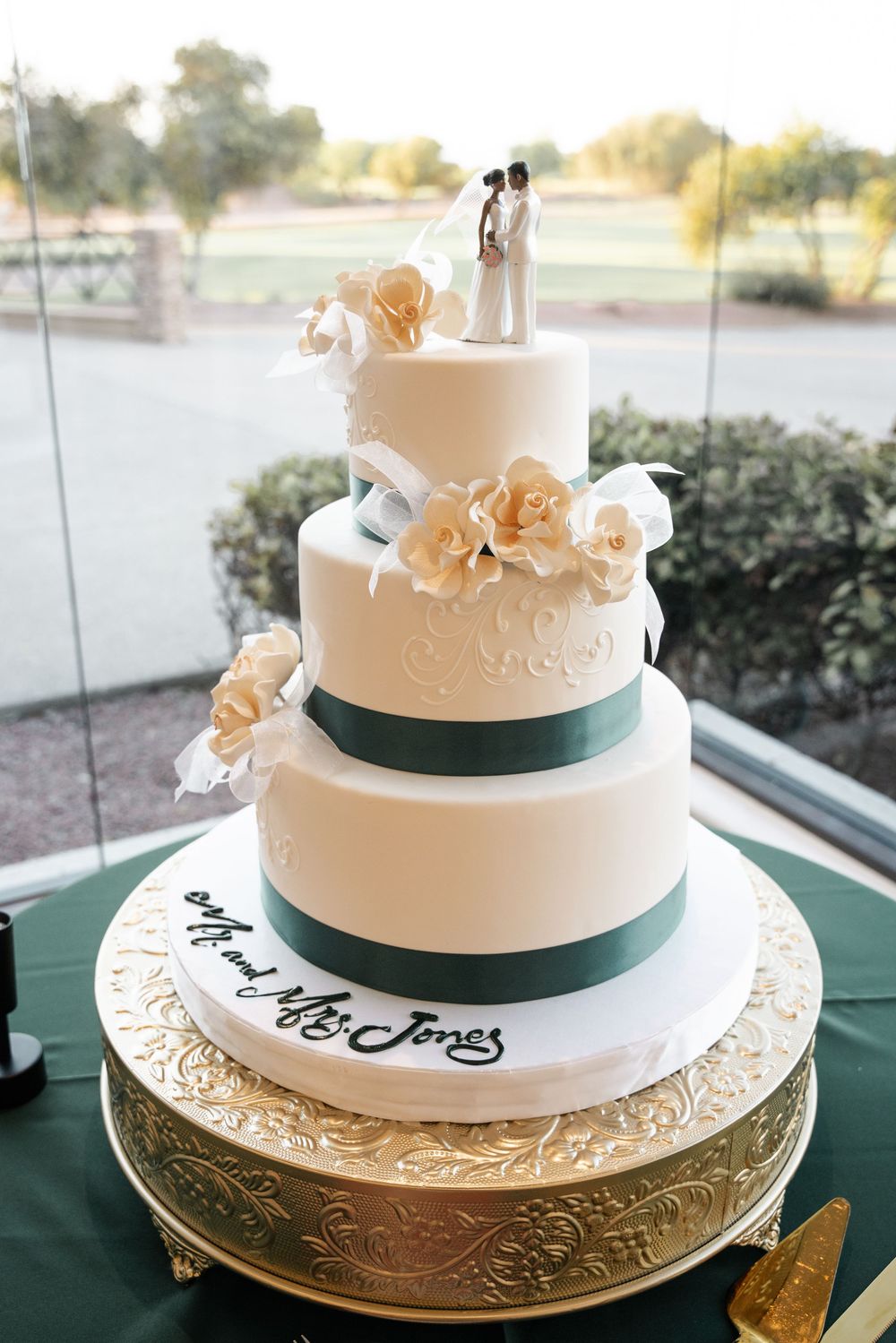 Elegant 4-tier white wedding cake with green ribbon, peach roses, and Mr. and Mrs. Jones inscription on gold stand.