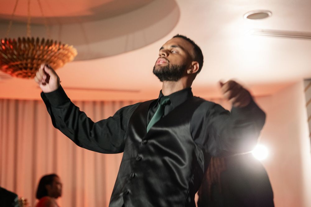 A man in a black vest and tie dances joyfully with arms raised at an elegant indoor event.