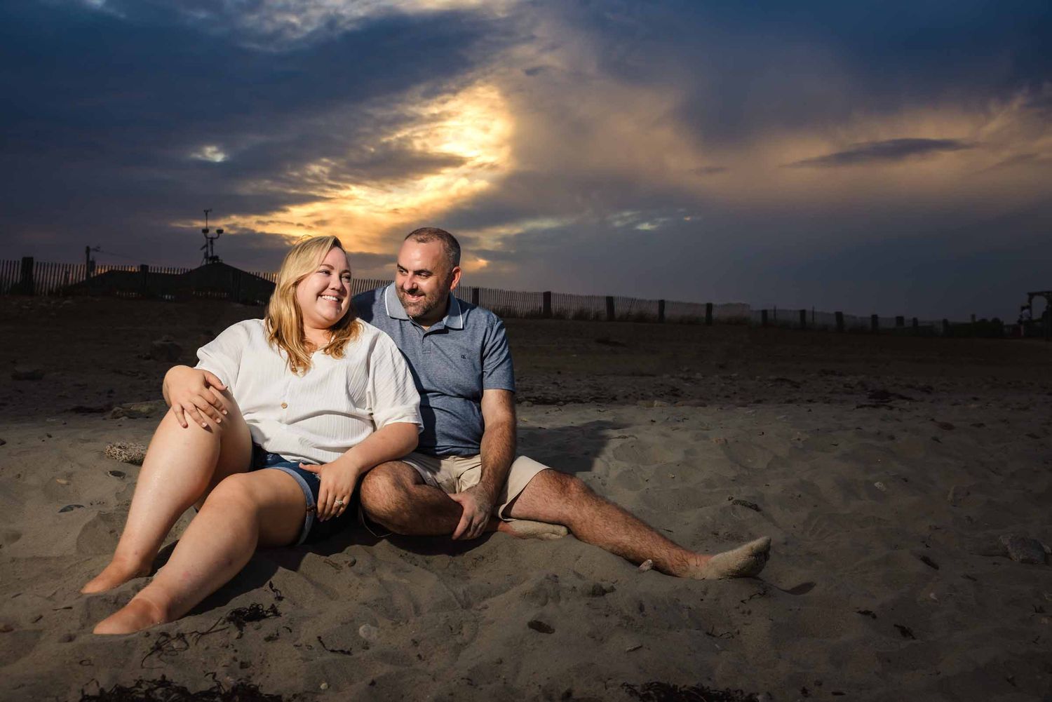 Engaged couple sitting together on the sand at Duxbury Beach, Massachusetts, laughing beneath a colorful dusk sky.