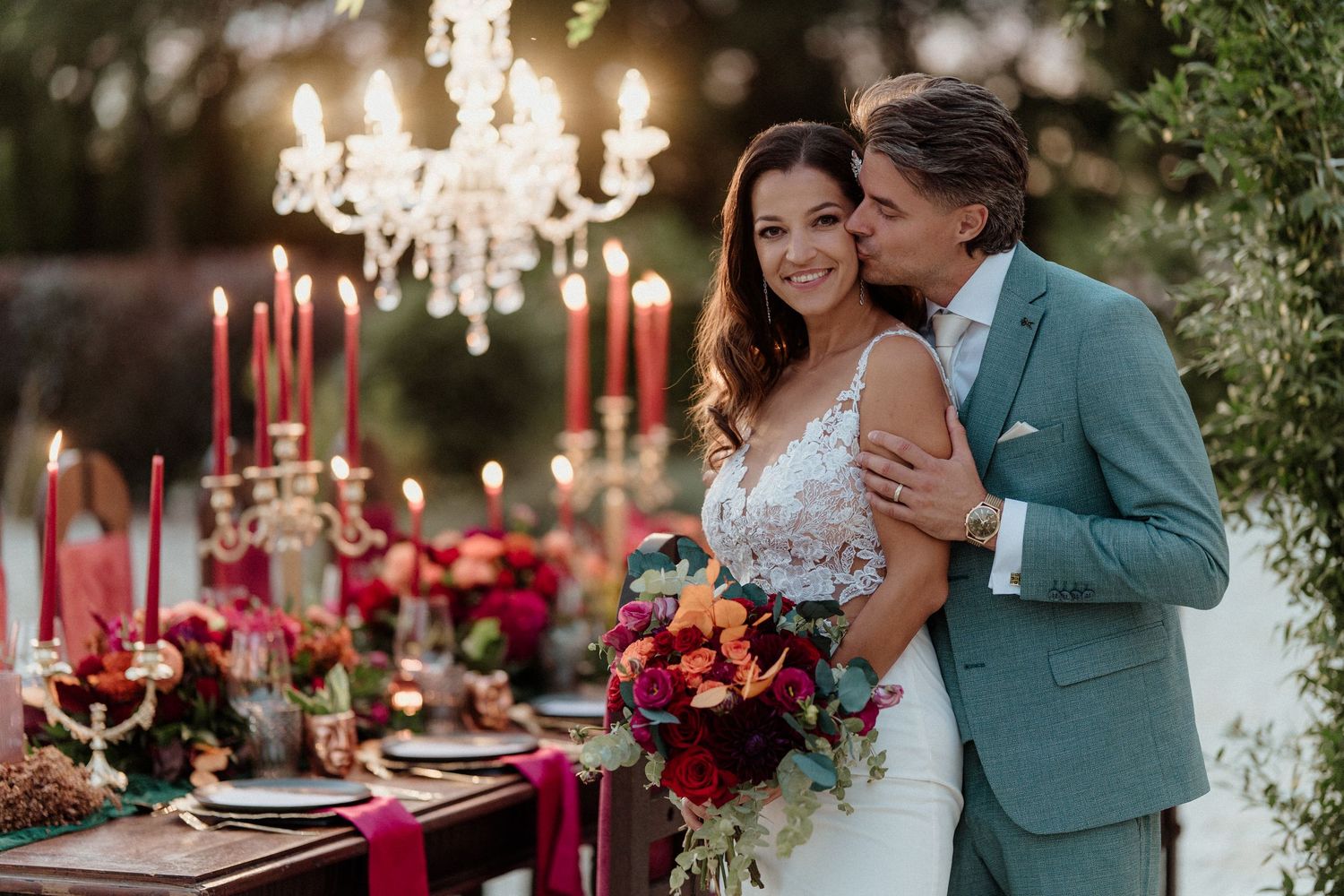 Couple embracing at decorated outdoor wedding table with candles and chandelier at dusk.