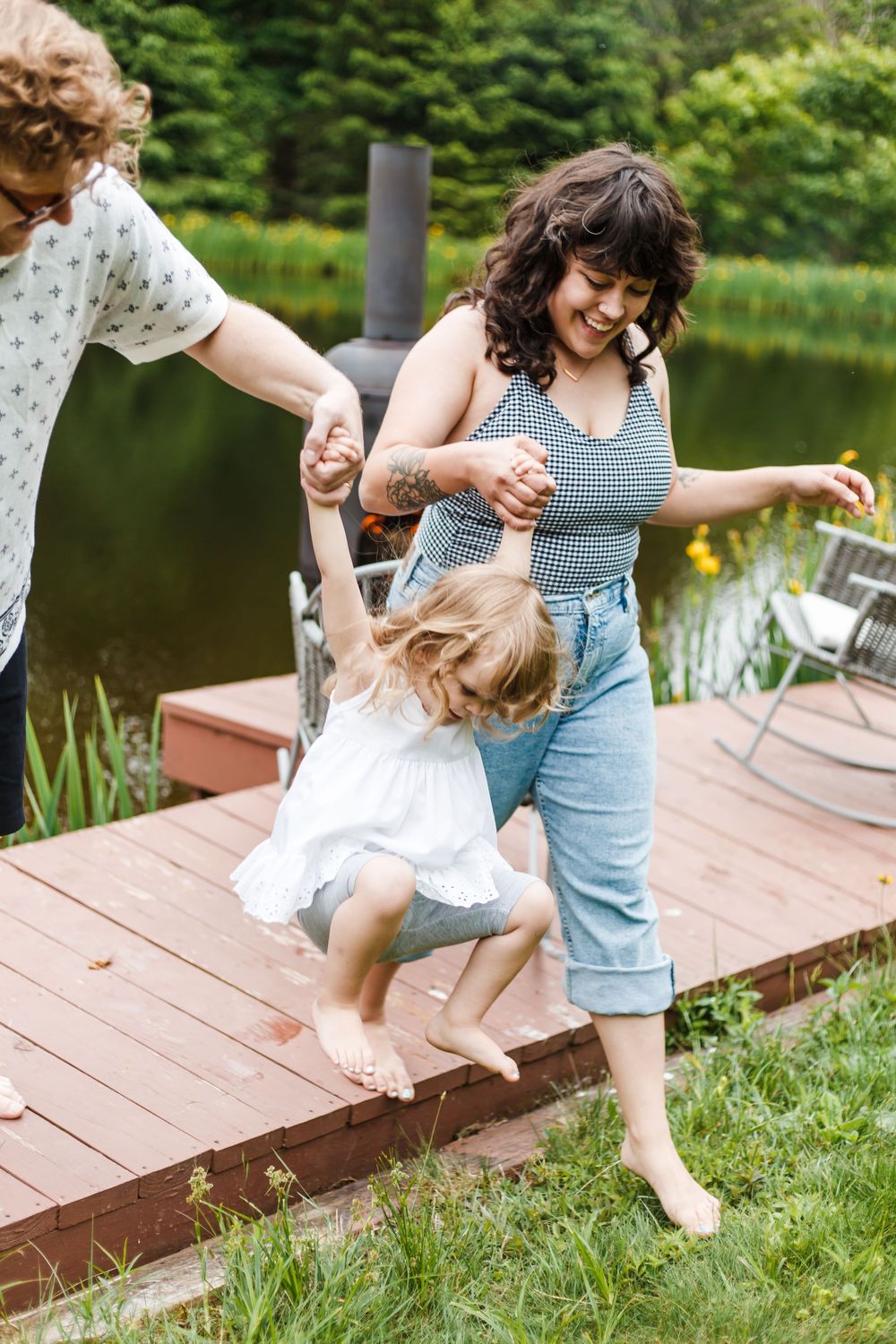 Family playing on a wooden deck by a pond, with a woman and man helping a young girl jump off the edge.
