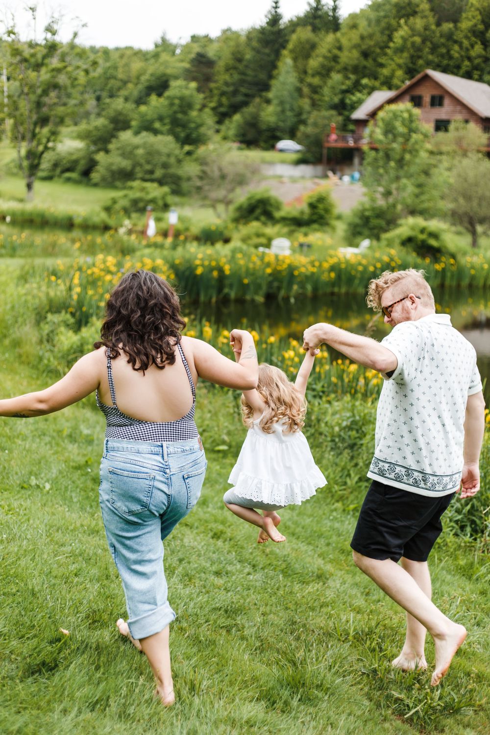 Family of three walking barefoot on lush green grass near a pond with yellow flowers and a rustic house.
