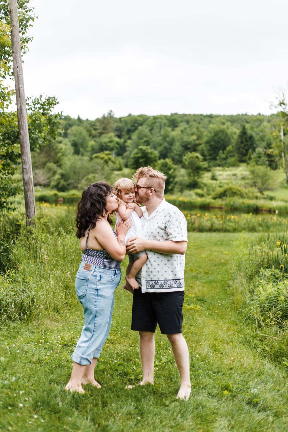 A young barefoot family shares a tender moment outdoors, parents kissing their toddler on a lush green grassy path.