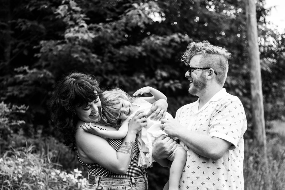 Black and white photo of a couple with glasses sharing a joyful moment with their young child outdoors.