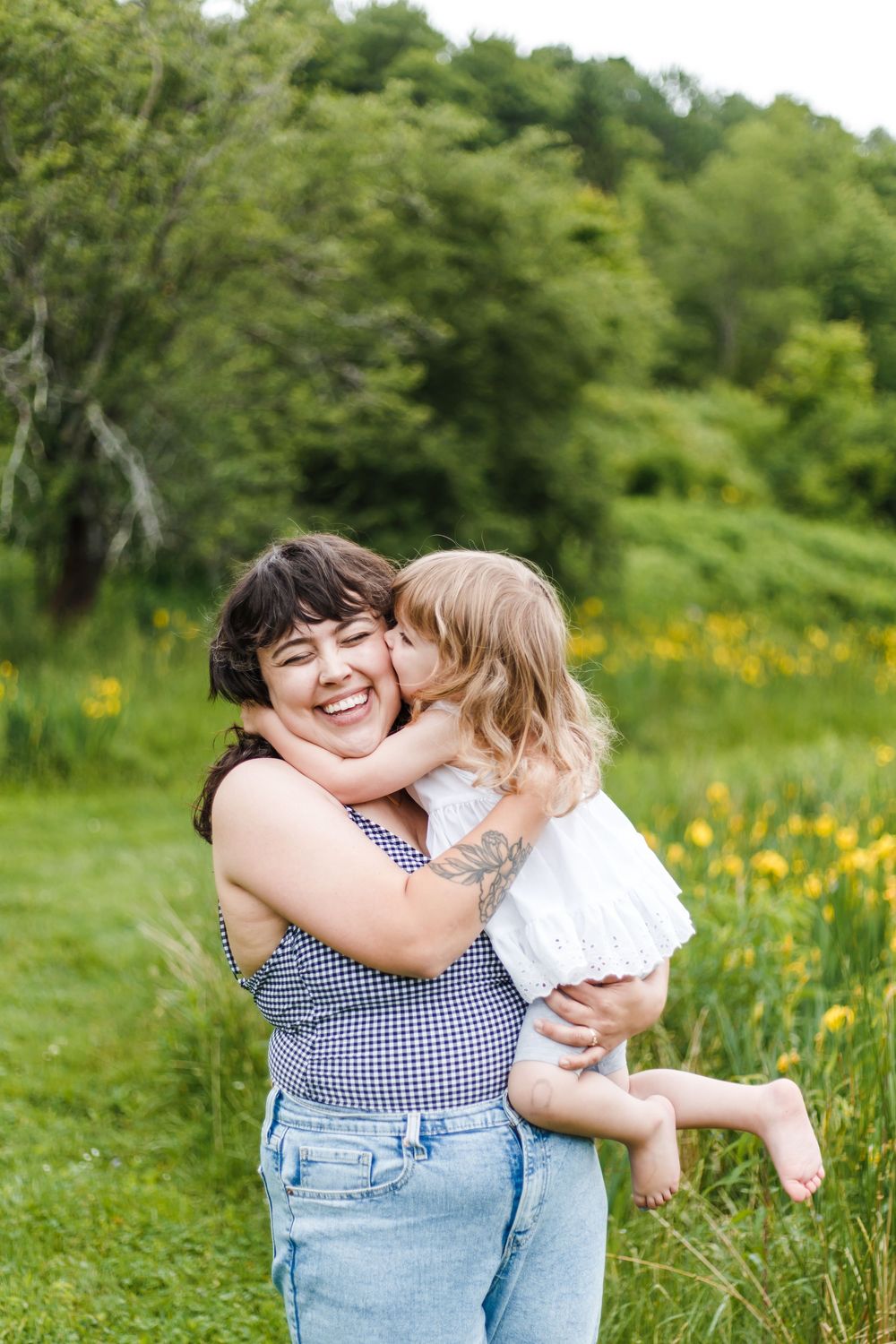 A laughing mother holds her young daughter in a sunny green meadow with yellow wildflowers as the child kisses her cheek.
