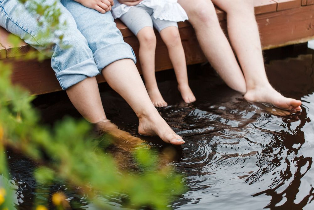 Barefoot family sitting on a wooden dock with feet dangling in calm water, surrounded by lush green foliage.