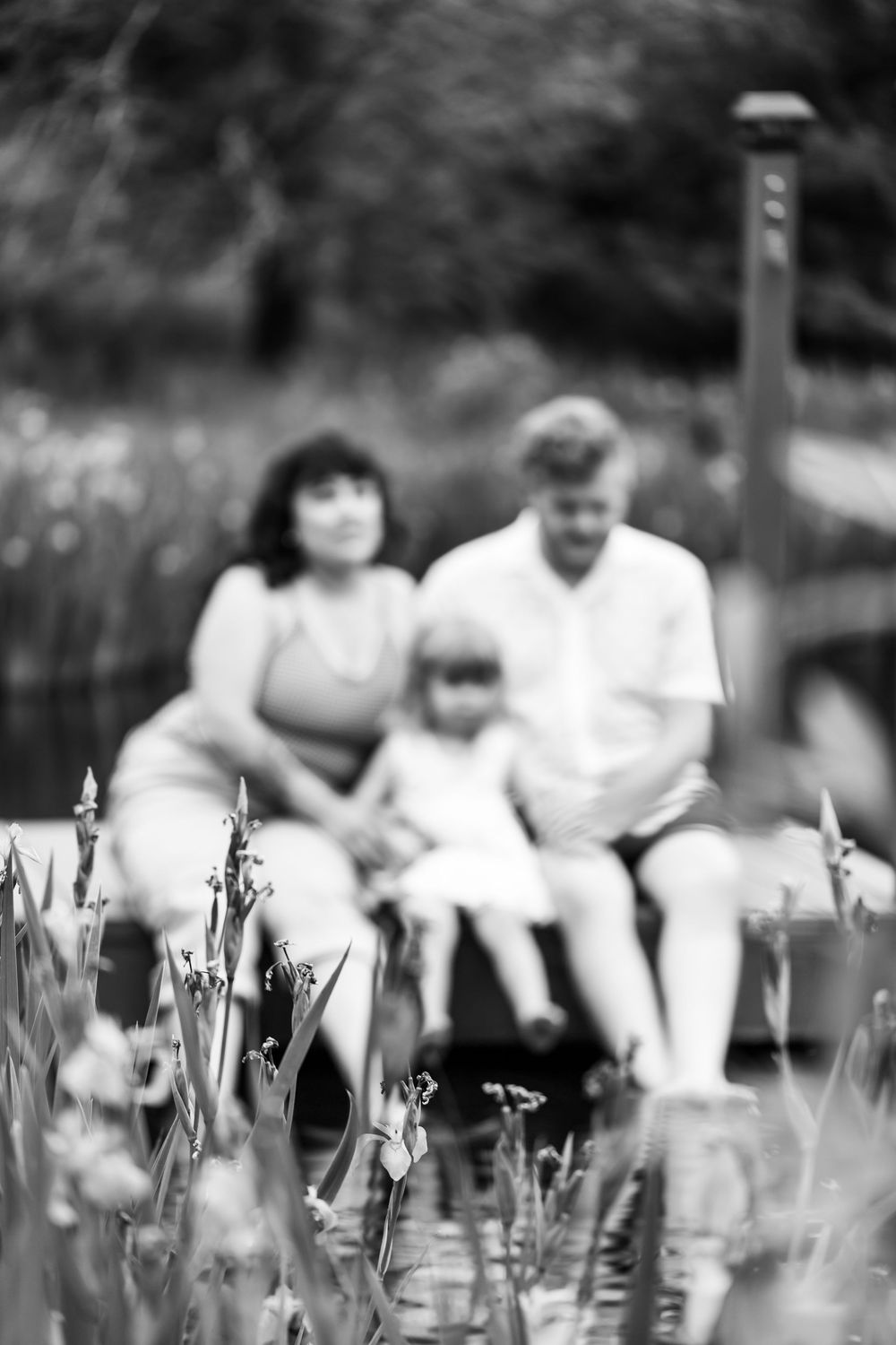 Black and white photo of a family of three sitting on a dock by a pond, surrounded by iris flowers in the foreground.