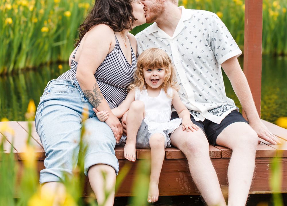 A pregnant woman and man kiss while their excited toddler sits between them on a wooden dock near a pond.