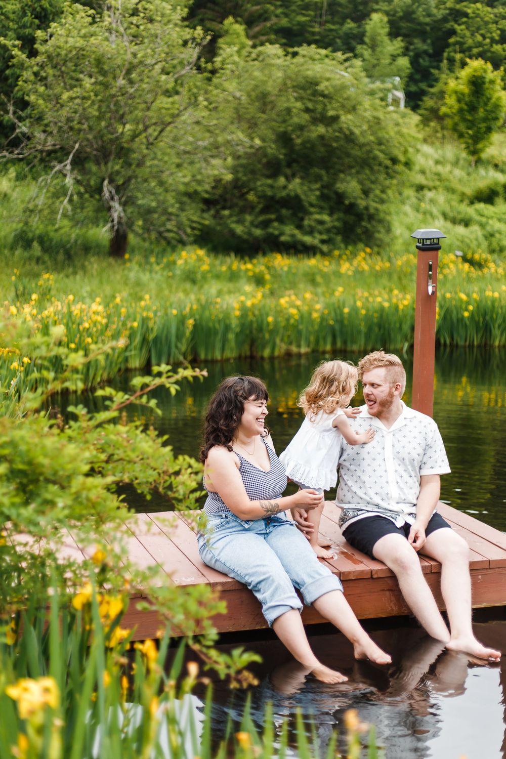 Happy family with young child sitting on a wooden dock by a scenic pond with yellow wildflowers in background.