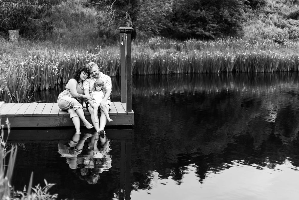 Family sitting together on a wooden dock over a calm lake, sharing a tender moment in black and white photography.