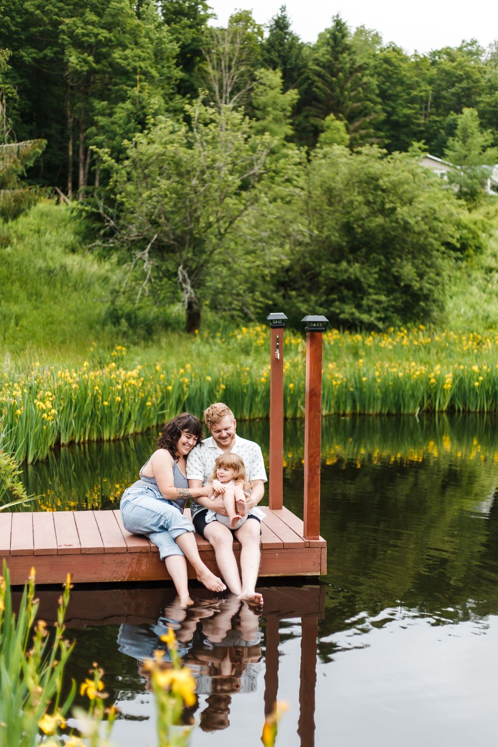 Happy family of three sitting on a wooden dock by a serene pond surrounded by lush greenery.