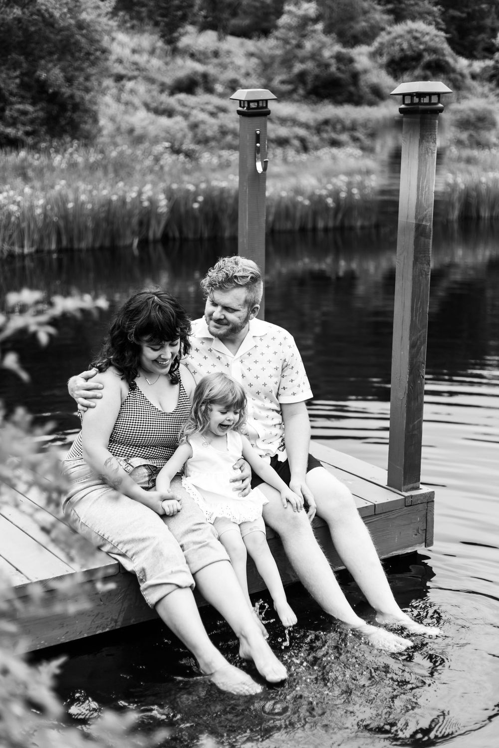 Black and white family photo of parents and child sitting on a dock with feet in the water.