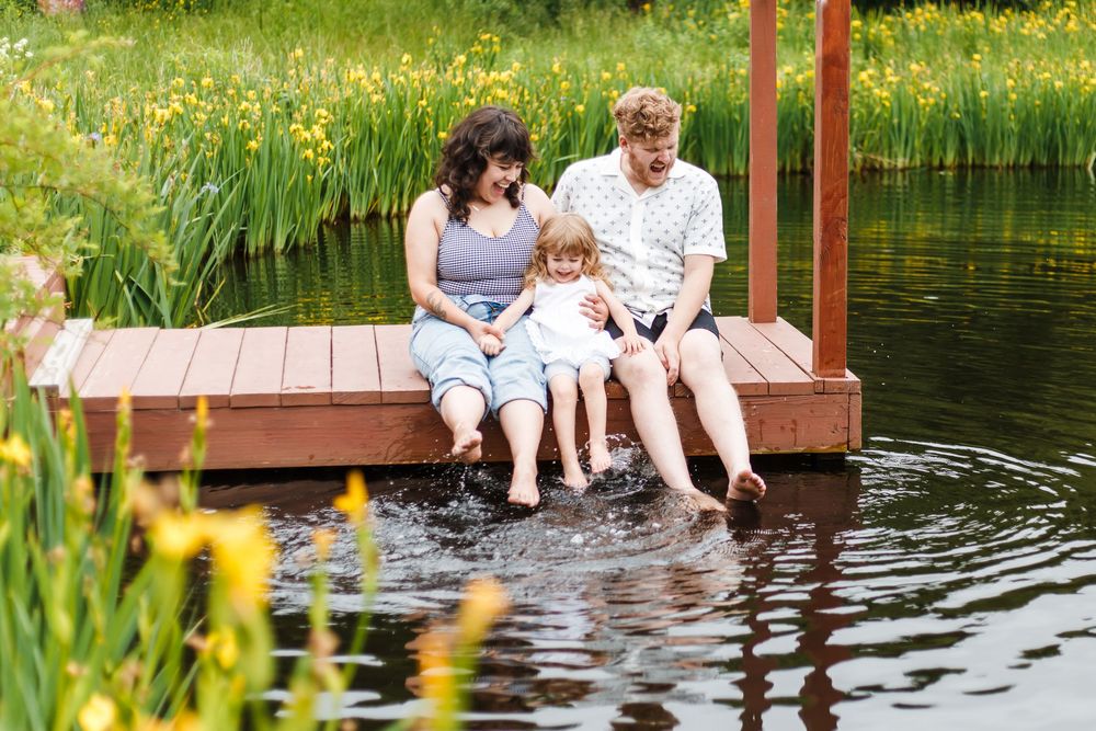 A family of three sits on a wooden dock, splashing their feet in a pond surrounded by lush greenery and yellow flowers.