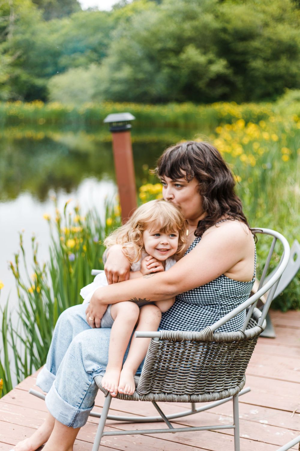 A mother sits in a wicker chair on a wooden dock, lovingly kissing her toddler daughter by a serene pond.