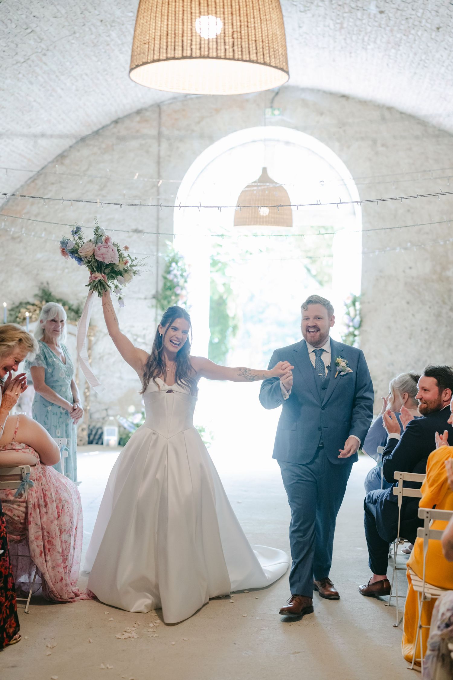 Bride raises bouquet joyfully as couple walks down aisle in stone arch wedding venue.