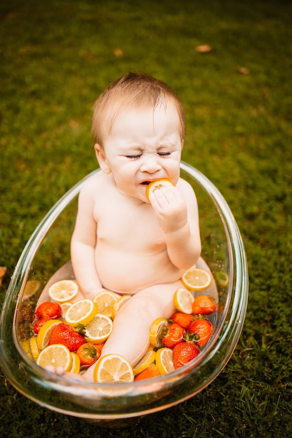 Baby sitting in glass bowl with strawberries and lemon slices outdoors, eating a lemon slice on green grass in Oak Ridge, TN.
