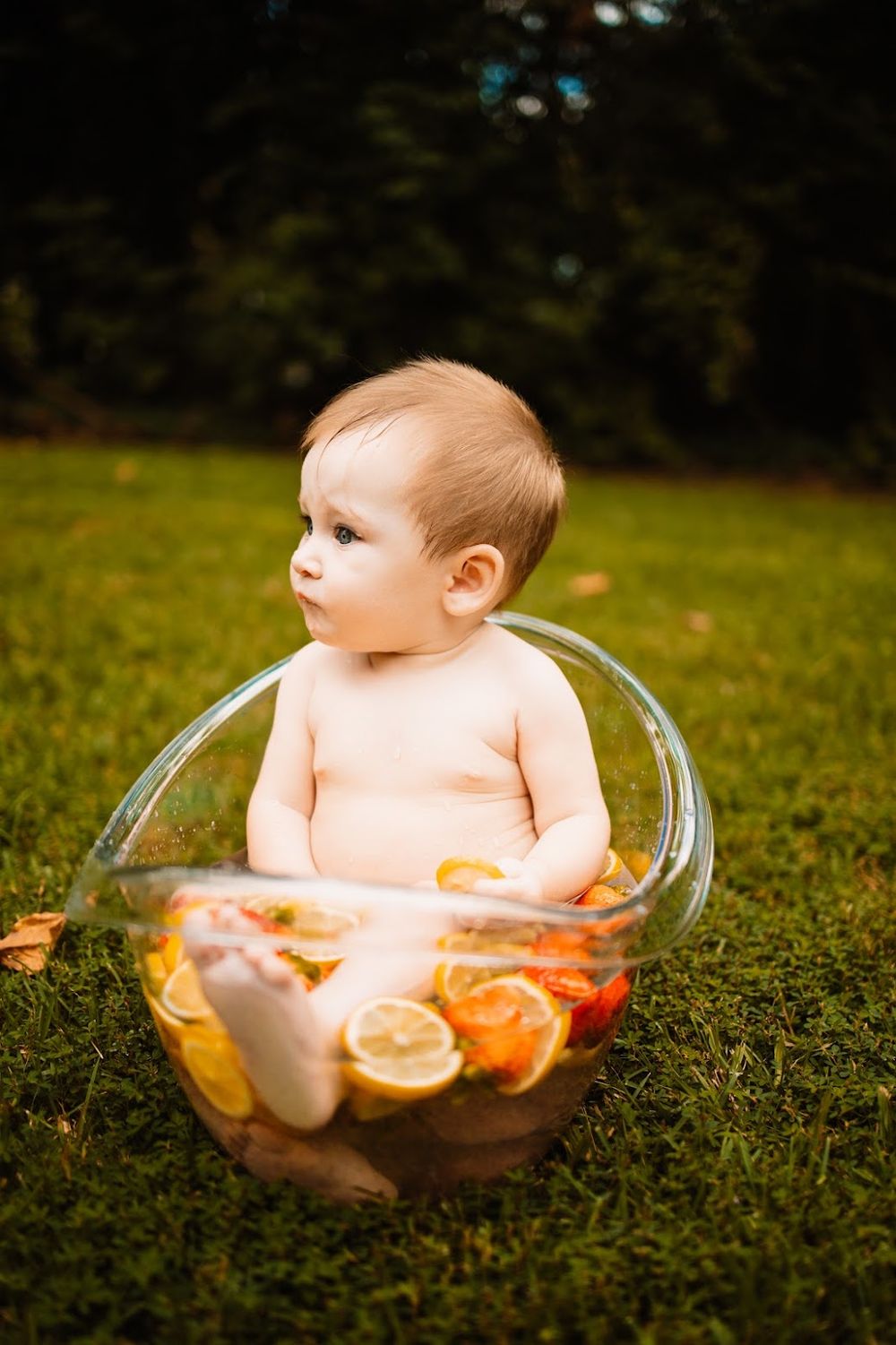 Cute baby sitting in a glass bowl filled with water and citrus fruit slices outdoors on green grass in Oak Ridge, TN.
