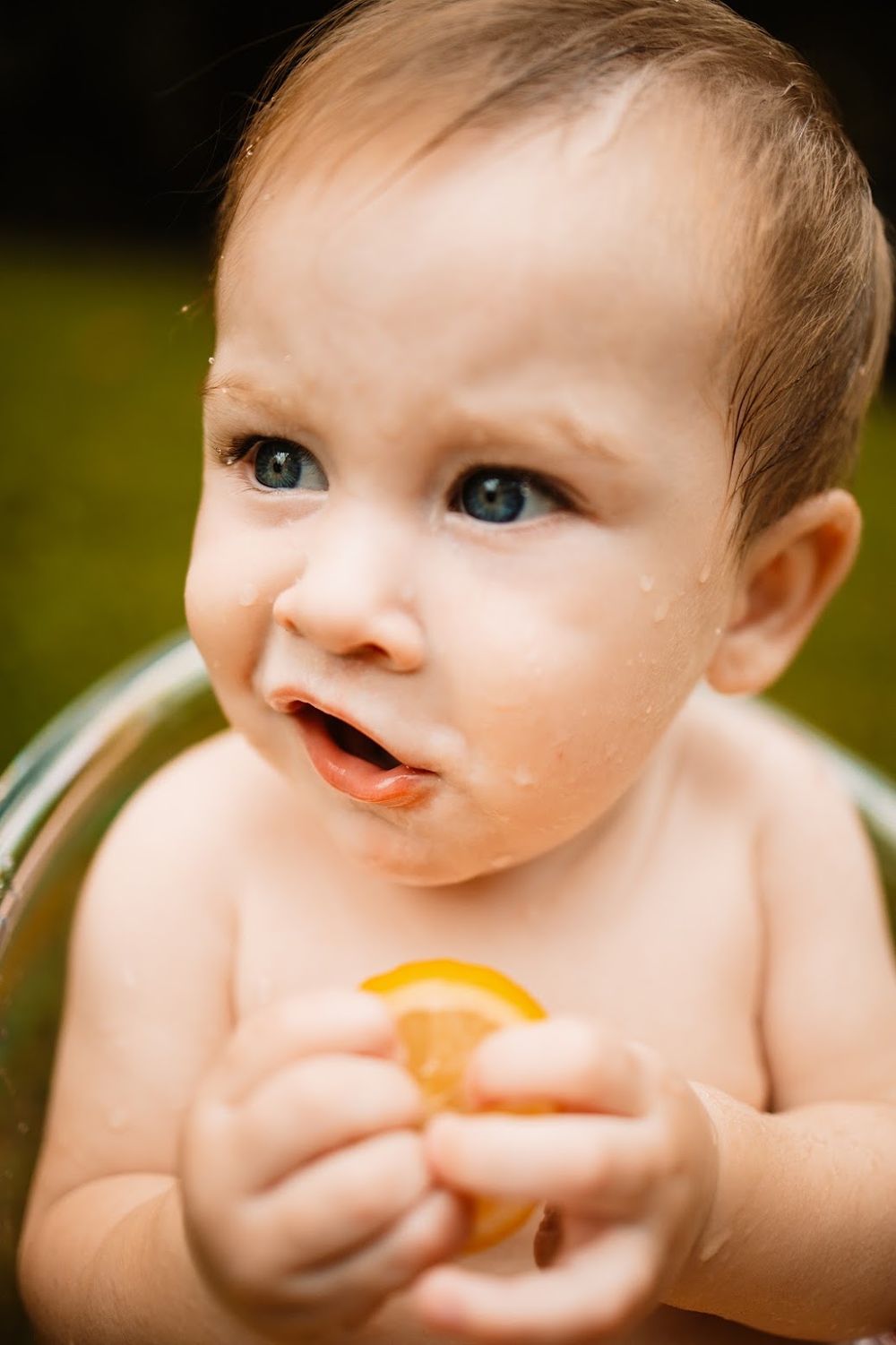 Adorable baby with blue eyes holding an orange slice in a fruit-filled tub  in Oak Ridge, TN.