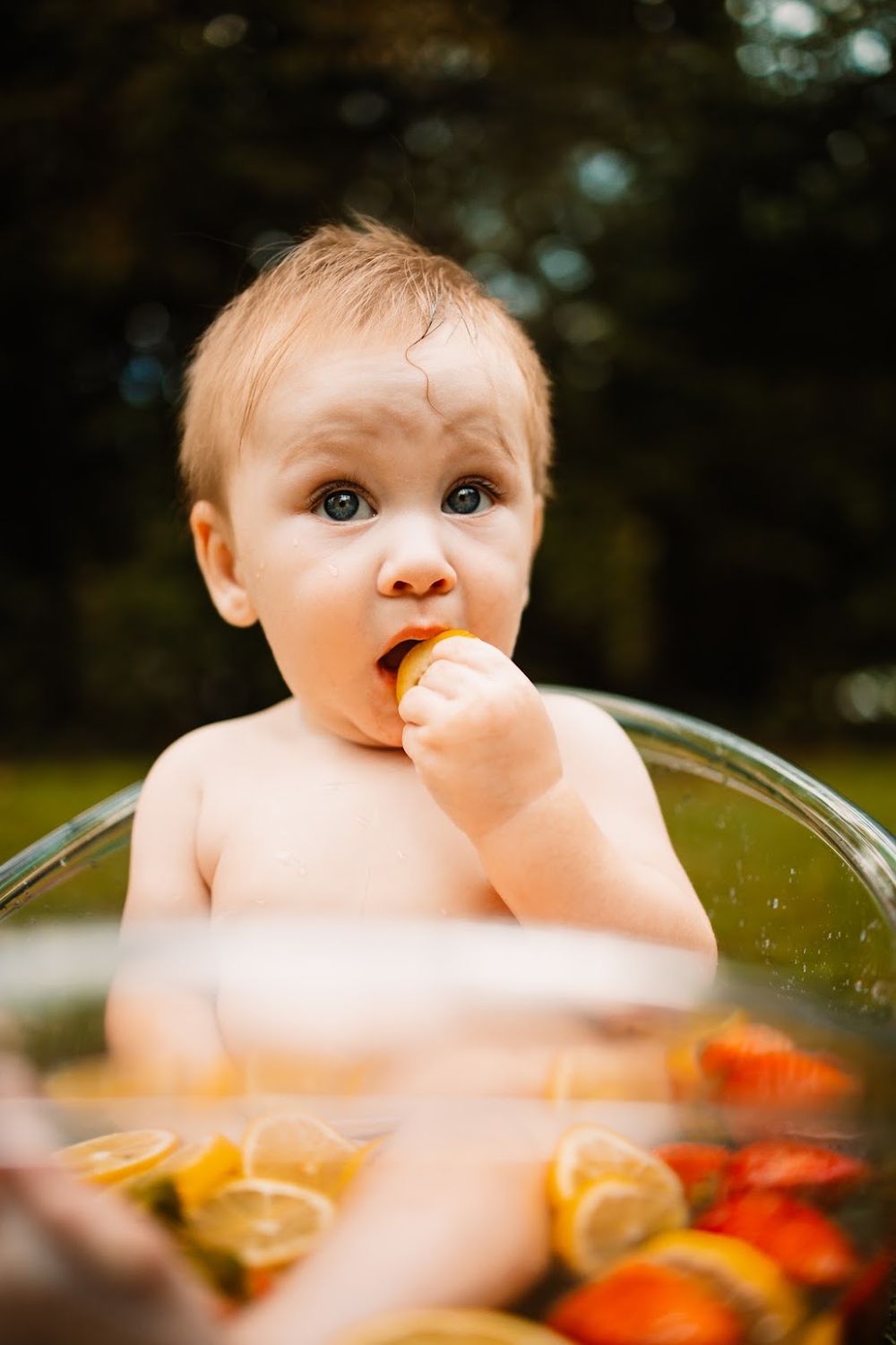 A curious baby sits in a clear tub filled with citrus fruits, nibbling on an orange slice outdoors in Oak Ridge, TN.