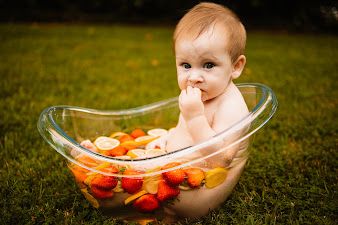 Cute baby sitting in a clear glass tub filled with colorful fruits outdoors on green grass  in Oak Ridge, TN.