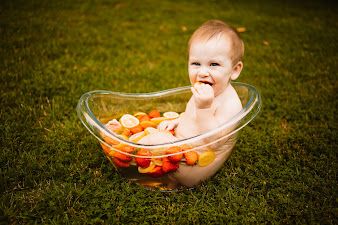 Baby sitting in a clear bowl filled with colorful fruits on green grass, smiling and enjoying a fun outdoor photo session  in Oak Ridge, TN.