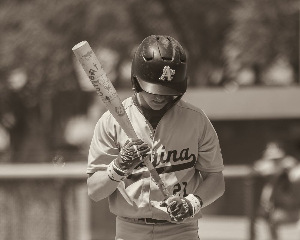 A baseball player in a Regina Athletics uniform examines his bat before an at-bat.