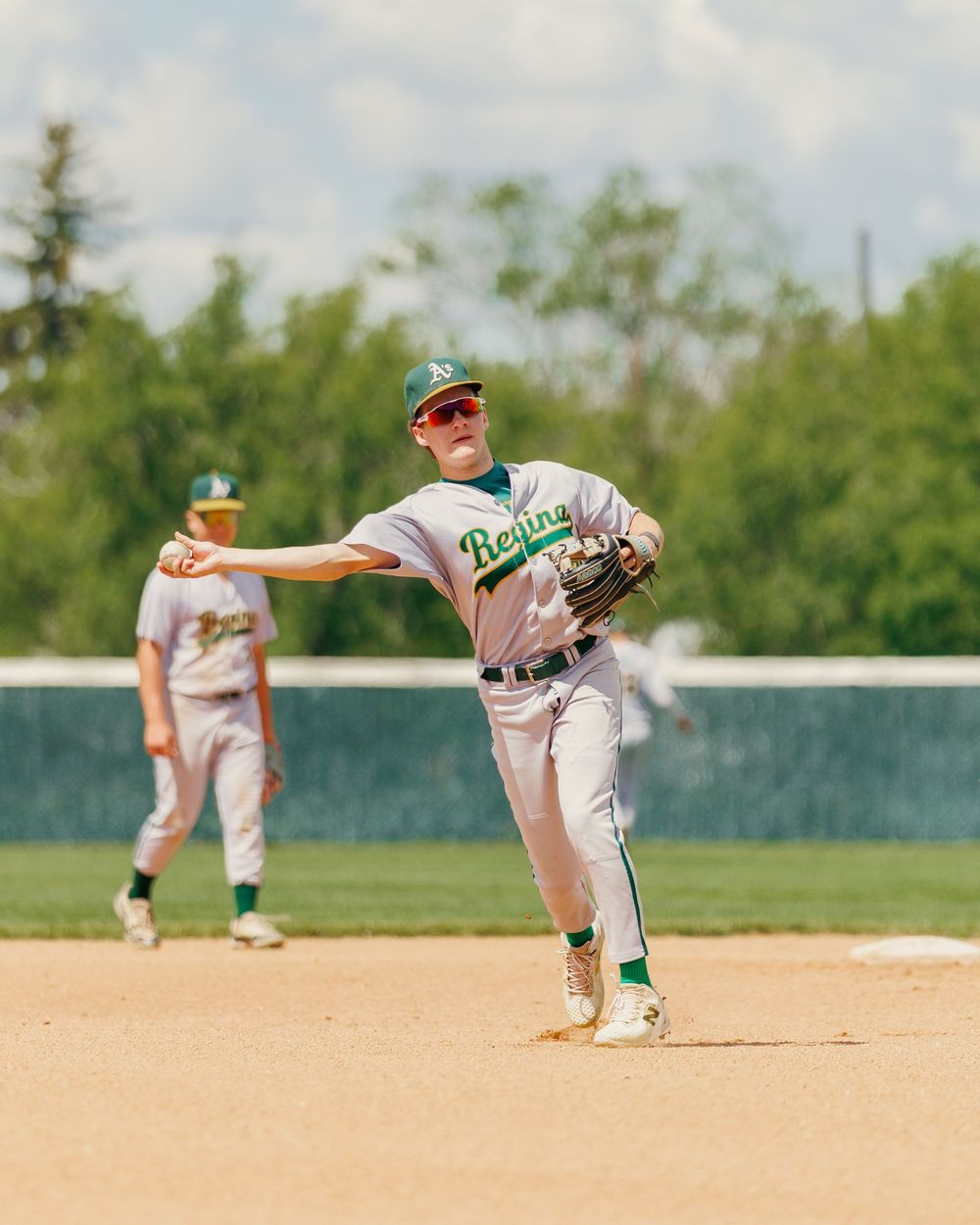 Regina baseball infielder in white and green uniform throws ball across diamond during sunny game.