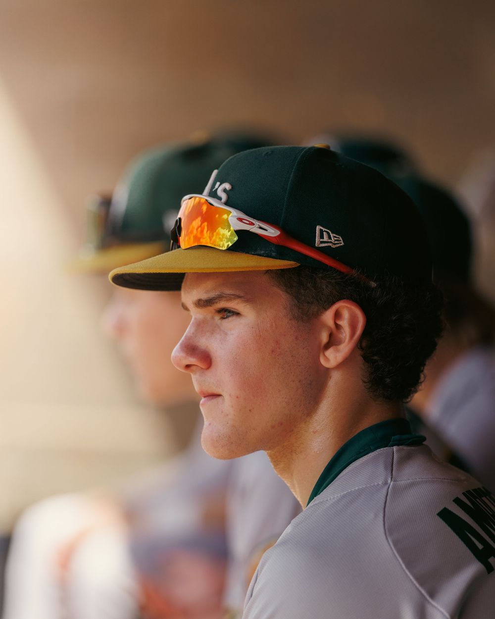 Young baseball player in green Regina cap and sunglasses watches game intently from the dugout.