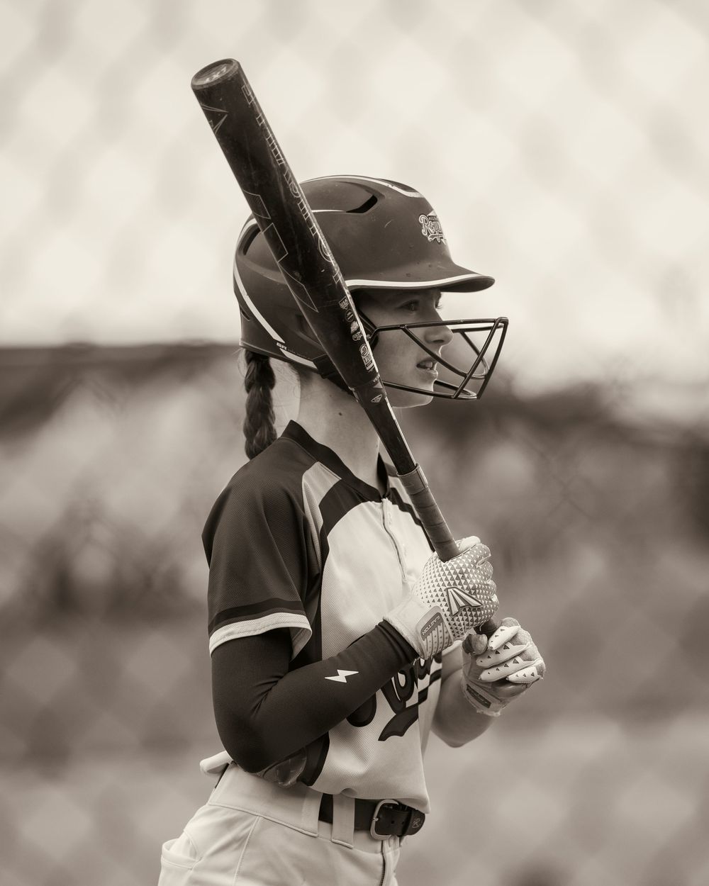 A softball batter in a dark uniform holds her bat ready in a sepia-toned photo.