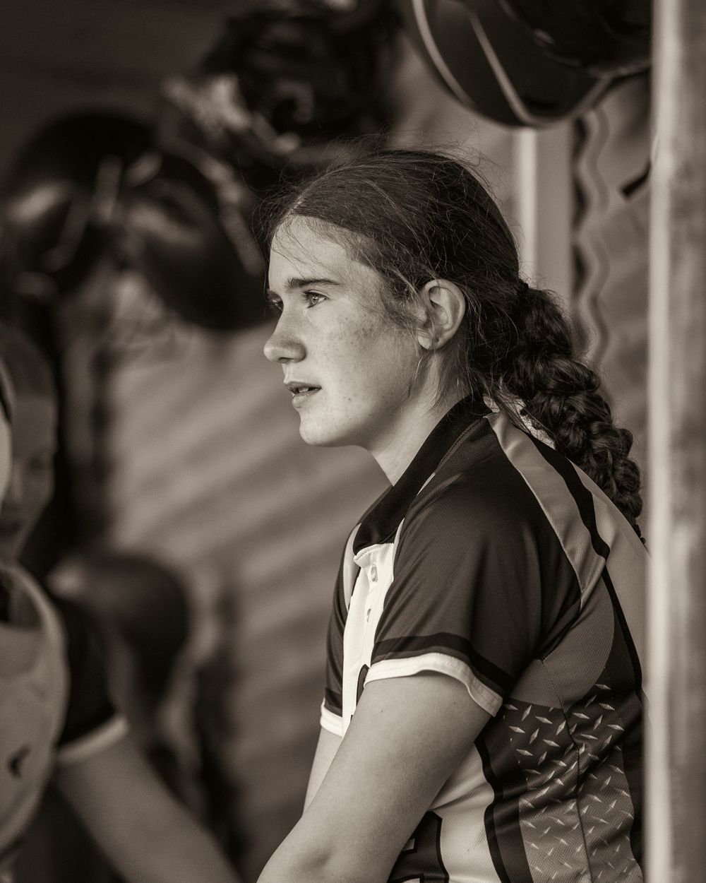A focused young softball player in catcher gear watches the game from the dugout.