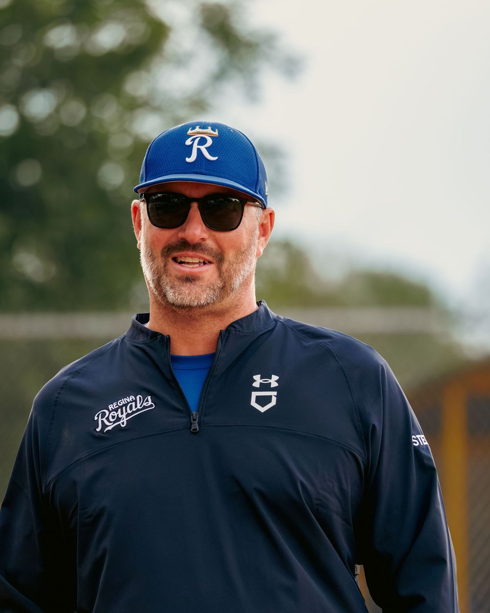 A smiling male softball coach wearing a Royals cap and sunglasses stands on the field.