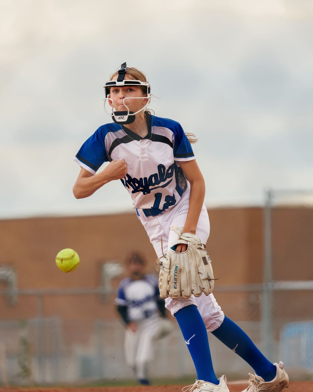 A Royals softball pitcher winds up to throw during a competitive game under cloudy skies.