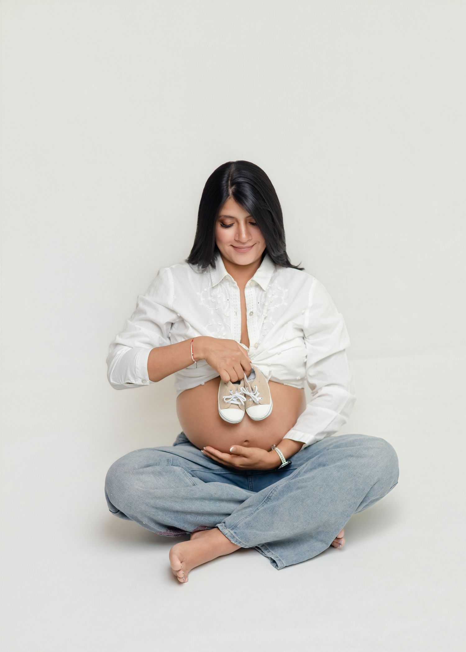 Pregnant woman in white shirt and jeans sitting cross-legged, holding tiny baby shoes on her bare belly.