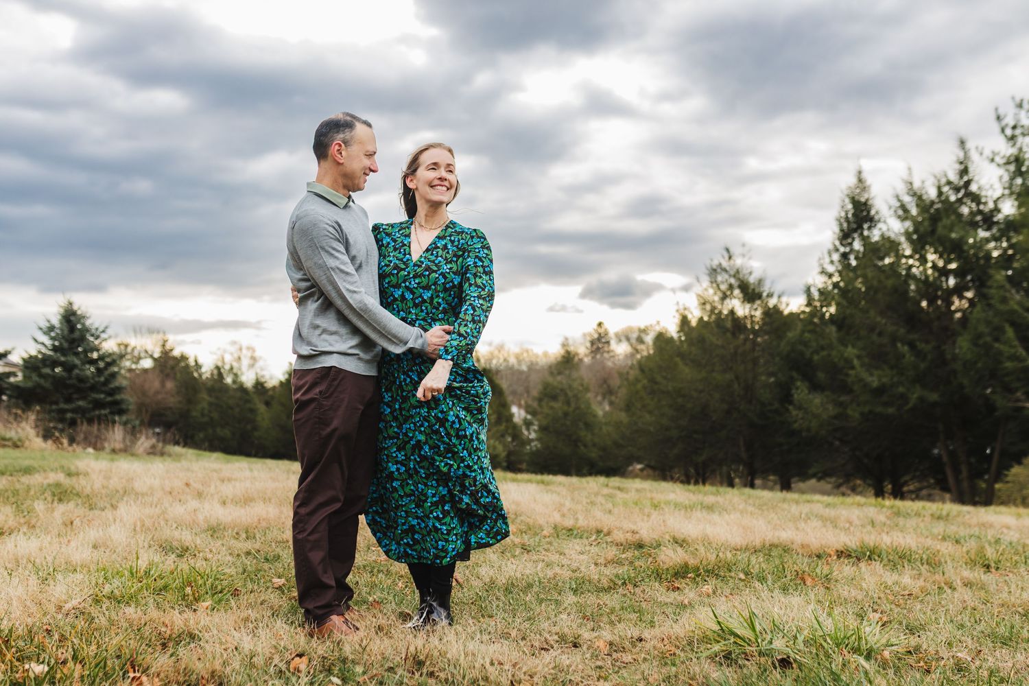 Couple laughing and holding hands in a grassy field with evergreen trees and cloudy sky backdrop.