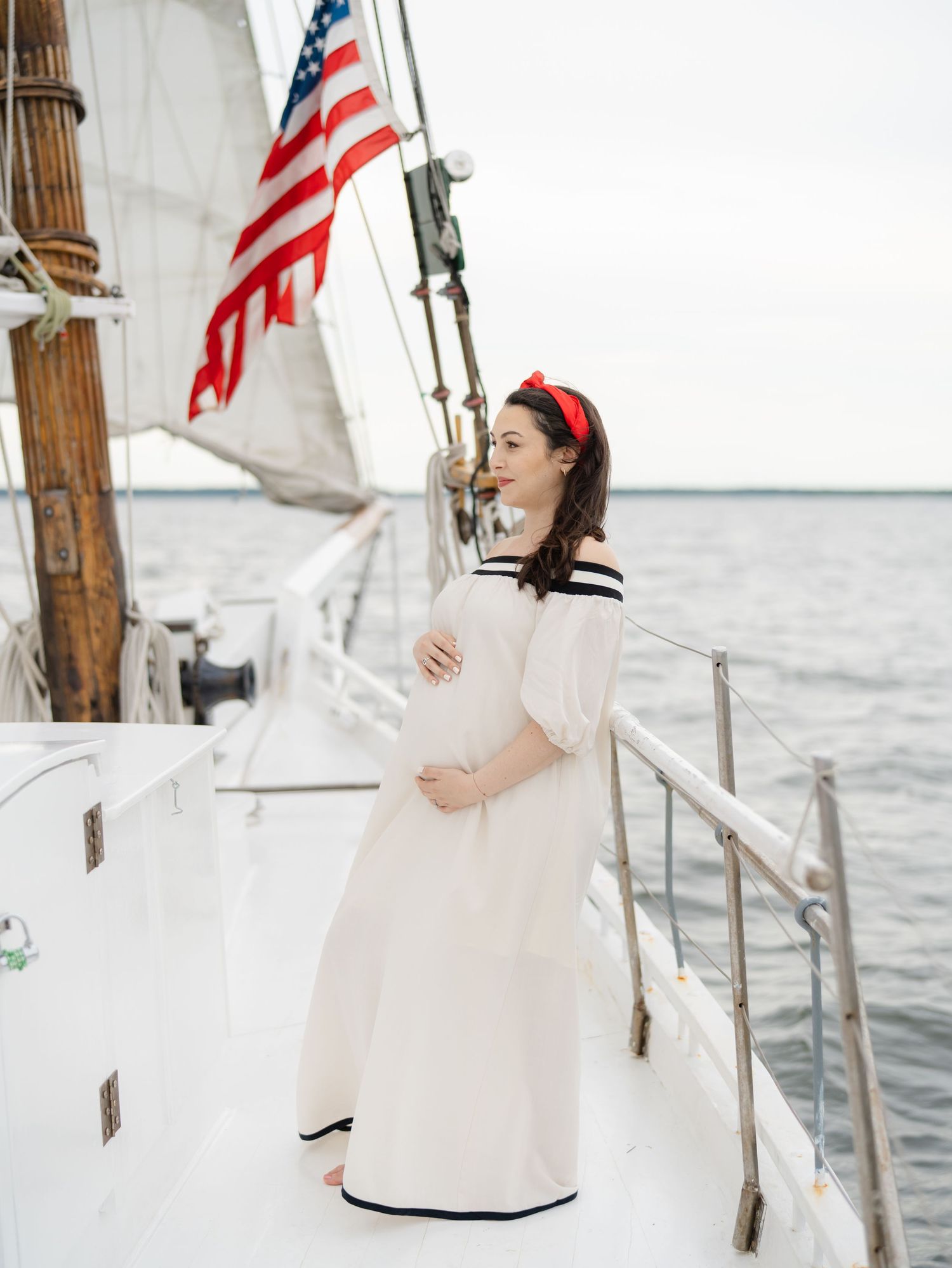 Belmar NJ photographer captures pregnant mother on a sailboat holding her belly in a white dress and red hair piece.