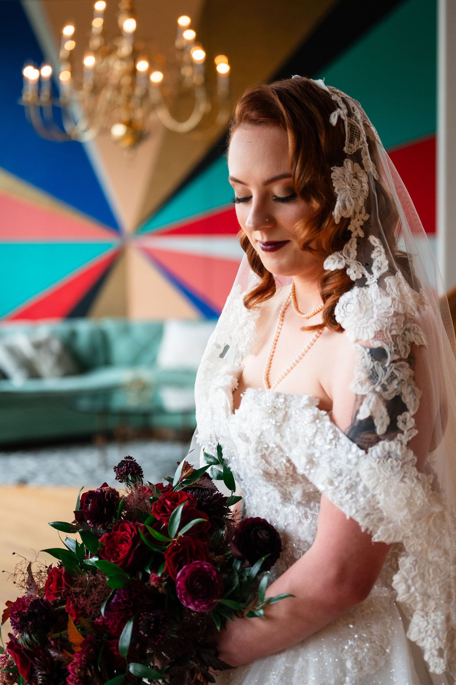 Bride in lace veil and white gown holds deep red bouquet, standing before colorful geometric wall with chandelier.