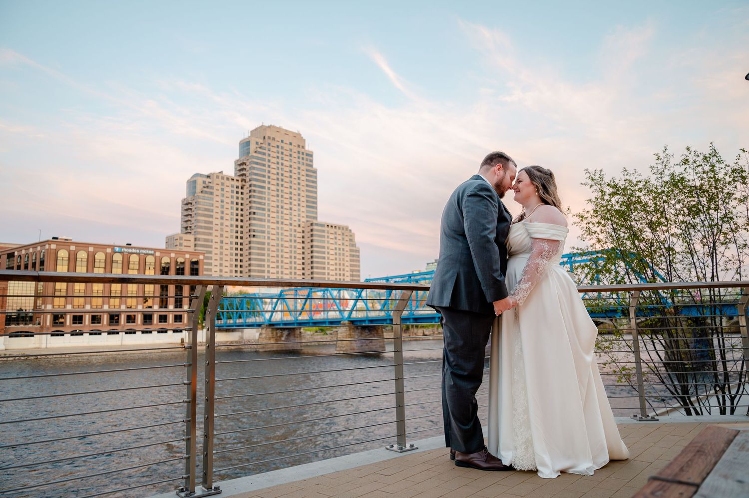 Bride and groom share a romantic kiss on a waterfront deck with a blue bridge and city skyline at sunset.
