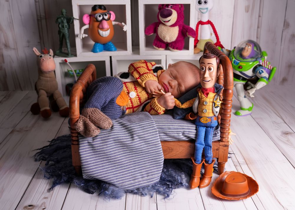 Newborn baby sleeping in tiny bed surrounded by Toy Story character dolls including Woody and Buzz Lightyear.