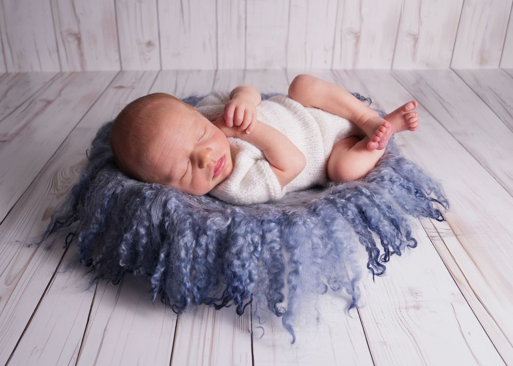Newborn baby sleeping peacefully in a blue fluffy bowl on a white wood floor during a professional photo shoot.