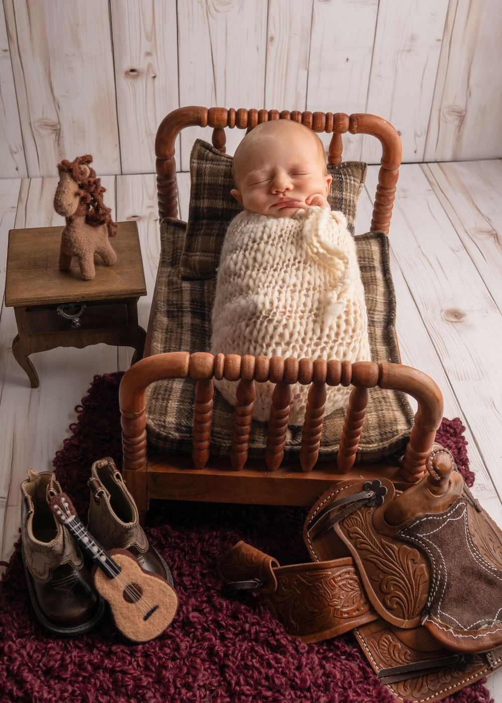 Newborn baby sleeping in miniature wooden bed with cowboy boots, saddle, ukulele, and stuffed giraffe props.