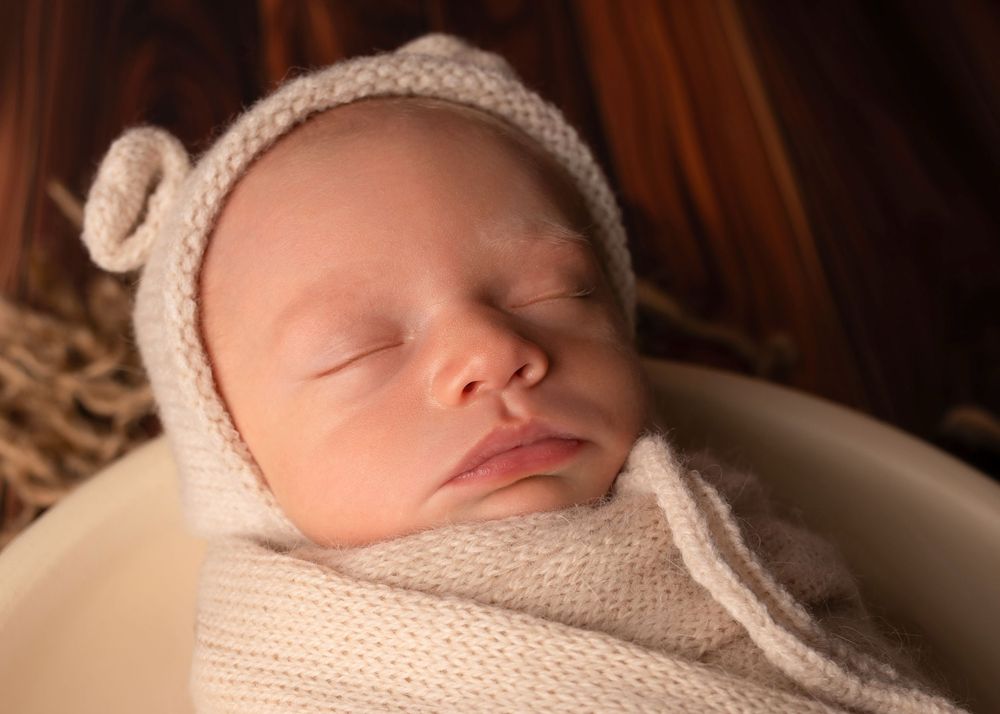 Peaceful newborn baby sleeping in a cream knit wrap and bear ear bonnet in a cozy bowl prop.