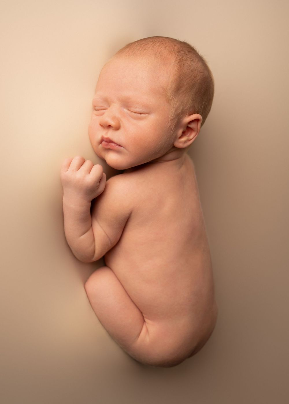 Peaceful sleeping newborn baby curled in fetal position on a warm beige background in a professional studio photo.