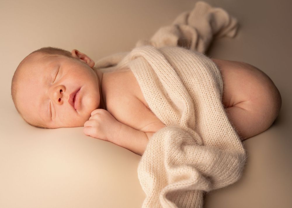 Peaceful newborn baby sleeping wrapped in a soft cream knit blanket on a neutral beige background.