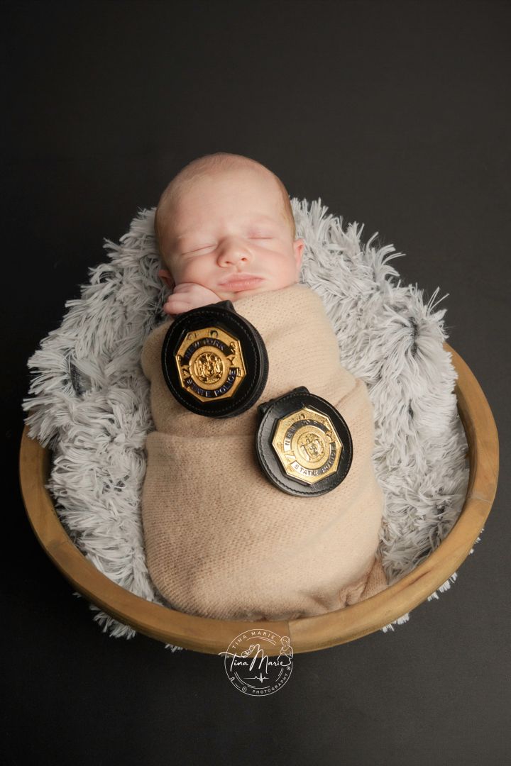 Newborn baby swaddled in beige wrap sleeping in wooden bowl with two gold law enforcement badges on dark background.