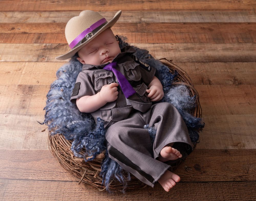 Newborn baby sleeping in a wicker basket wearing a tiny ranger hat, gray uniform, and purple tie on a wooden floor.