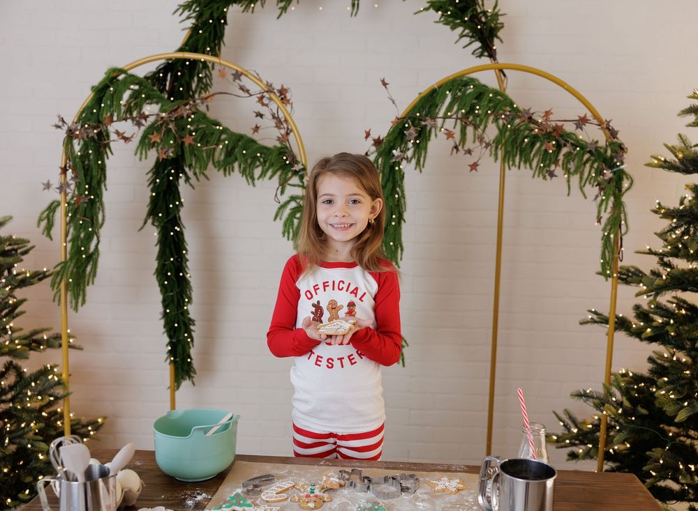 Young girl in Christmas pajamas smiling behind a baking table with holiday greenery arch backdrop.