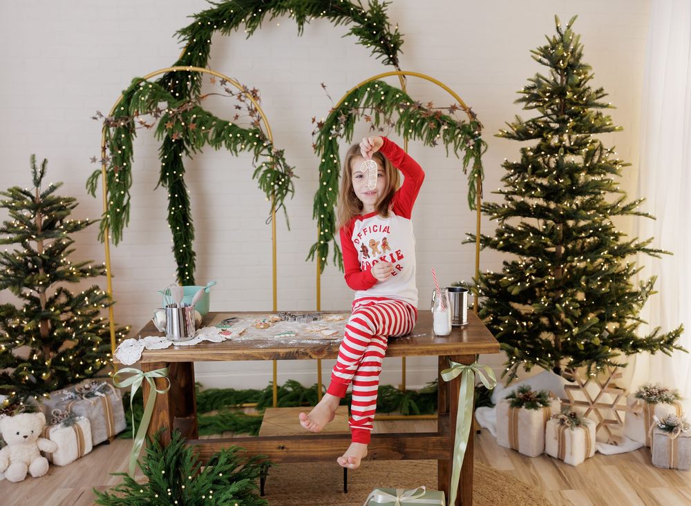Young girl in red and white striped Christmas pajamas sitting on a wooden table surrounded by festive holiday decor.