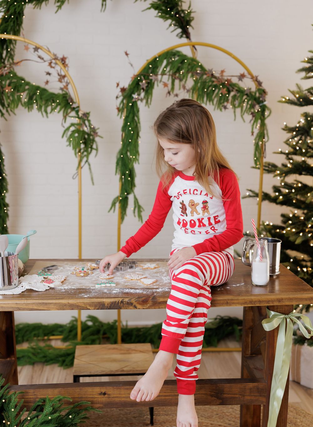 Girl in Christmas pajamas decorating cookies on a wooden table with holiday greenery and tree in background.